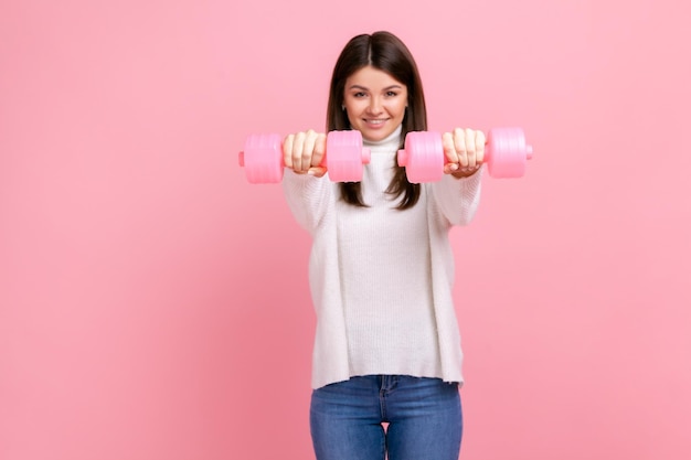 Young adult sporty woman with dark hair holding out pink dumbbells looking at camera healthy lifestyle wearing white casual style sweater Indoor studio shot isolated on pink background