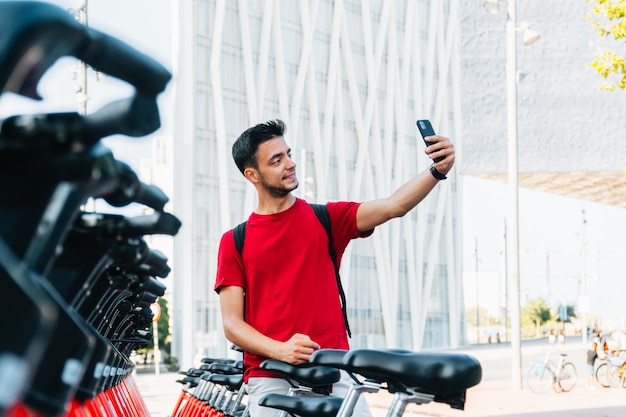 Foto giovane studente adulto che si fa un selfie con il cellulare in uno stand di noleggio biciclette