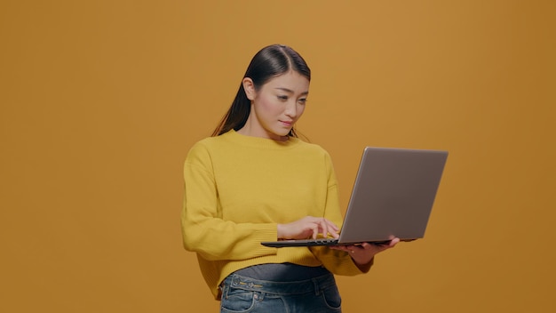 Young adult typing on laptop pc with technology in studio. Asian woman using portable computer to browse internet, advertising freelance work and modern communication on device.