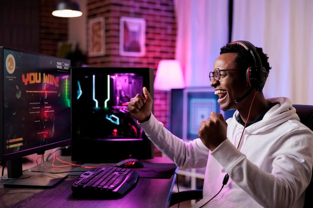 젊은 성인이 PC에서 온라인 비디오 게임 토너먼트를 우승하고 인터넷에서 여러 플레이어와 액션 게임 플레이를 스트리밍합니다. 사격 경쟁을 하고 승리에 대해 행복감을 느끼는 남성 게이머.