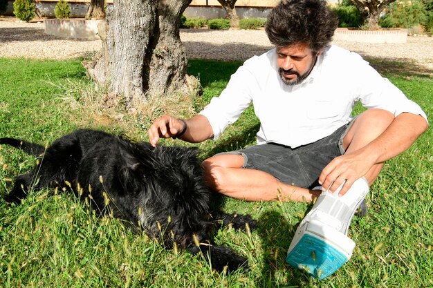 Young adult with a fractured foot and a walker boot playing\
with his pet, a black schnauzer dog,