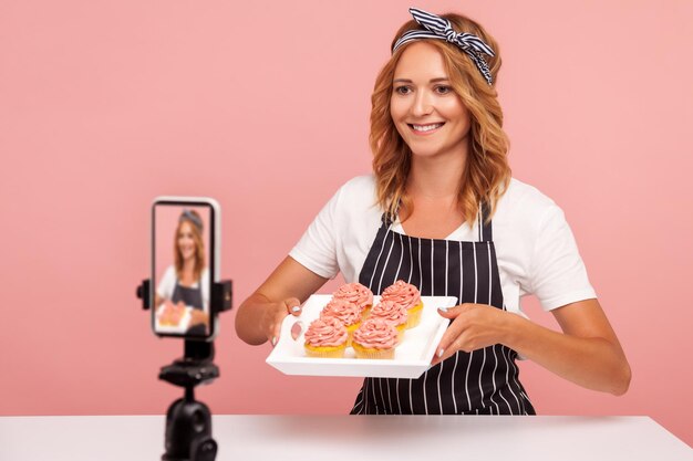 若い大人の女性ブロガーが焼きたてのケーキを見せてビデオで撮影し、フードチャンネルのビデオを録画し、女性のパティシエが携帯電話でぶらぶらしています。ピンクの背景に分離された屋内スタジオショット。