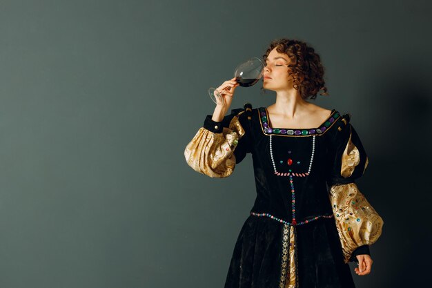 Young adult woman dressed in a medieval dress holding a glass of wine and drinking a red wine