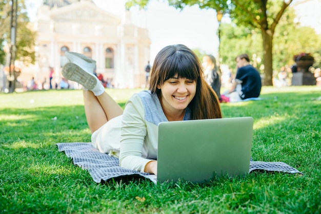 緑の芝生の上の都市公園でラップトップで横たわっている若い大人の女性