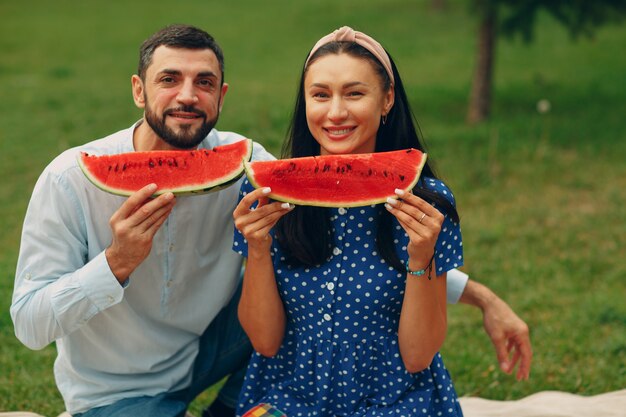 Young adult woman and man couple picnic at green grass meadow in park having fun and smile with watermelon