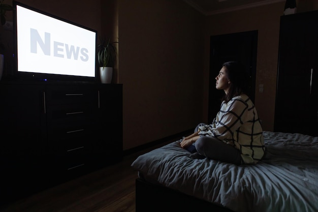 Young adult woman sitting on bed looking tv white screen copy\
space night time