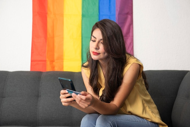 young adult woman sitting on the couch shopping online with her smartphone with a rainbow flag