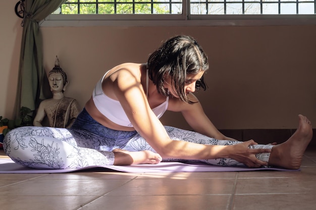 A young adult woman sitting on mat stretching leg at home