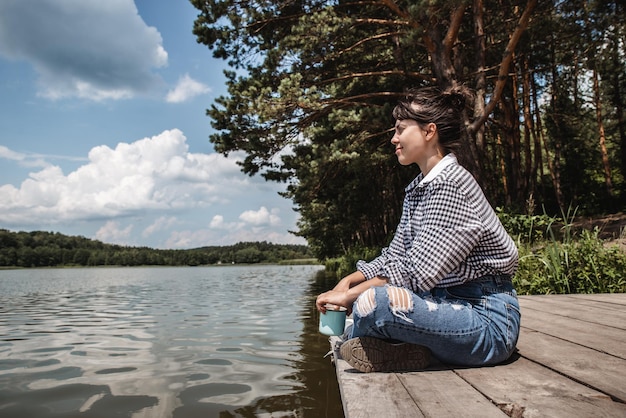 Young adult woman sitting on wooden dock drinking coffee and looking at lake