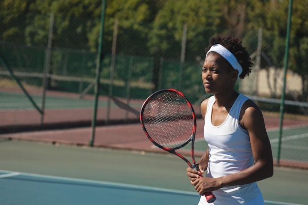 Young adult woman standing on outdoor tennis court gripping tennis racket with headband copy space