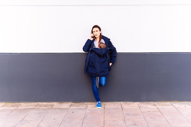 Young adult woman talking on the smartphone carrying her baby leaning on a gray and white wall with a serious gesture receiving bad news