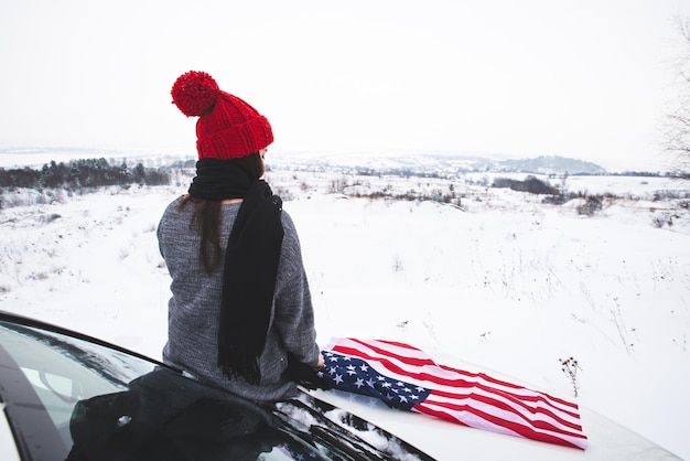 Young adult woman traveler sitting on the hood of the suv car with usa flag