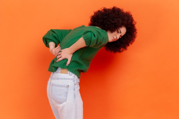 Photo young adult woman with afro hairstyle wearing green casual style sweater suffering from lower back pain, pinched nerve or kidney inflammation. indoor studio shot isolated on orange background.