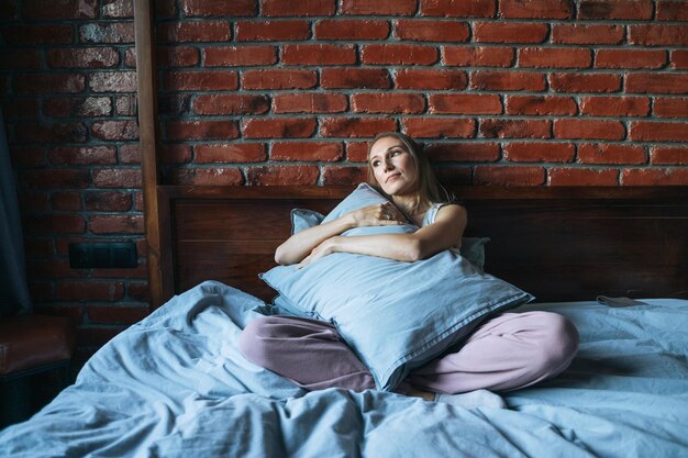 Young adult woman with blonde long hair in casual sitting on bed with pillow at home