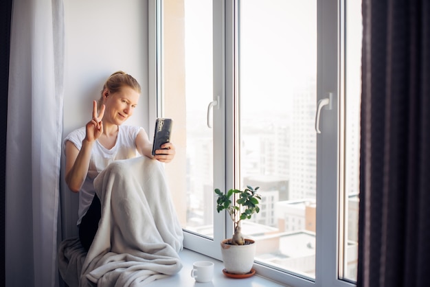 Young adult woman with glasses takes a selfie sitting on the windowsill at home, gesturing with her hand. Female smiles while chatting, looking at smartphone.