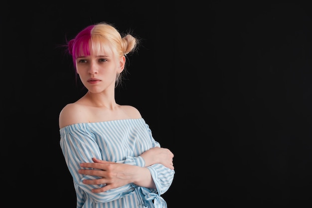 Young adult woman with pink and white hair hugging herself on black background