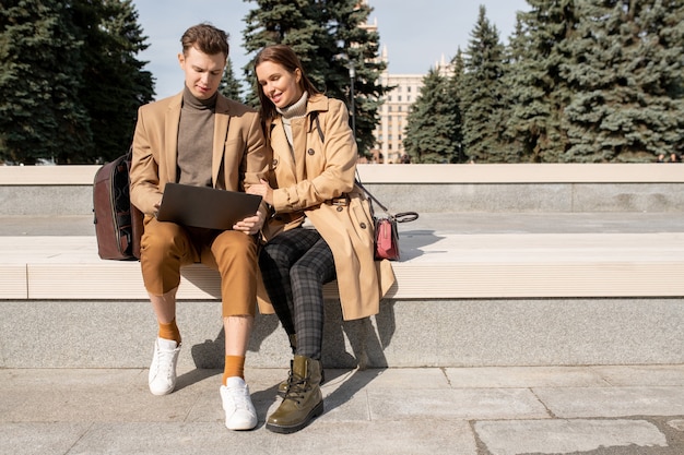 Молодая ласковая пара в элегантных бежевых плащах, пуловерах и брюках смотрит на дисплей ноутбука во время просмотра онлайн-видео на открытом воздухе