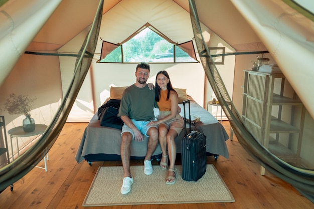 Young affectionate couple sitting on double bed inside glamping house