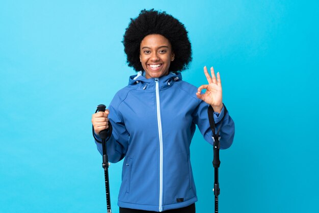 Young Africa American with backpack and trekking poles isolated on blue background showing ok sign with two hands