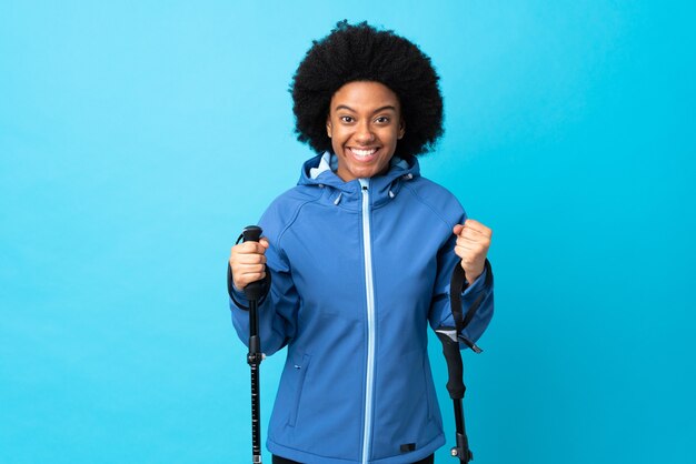 Young Africa American with backpack and trekking poles isolated on blue celebrating a victory in winner position