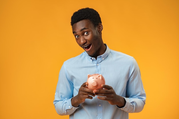 Young african amercian man holding piggy bank over yellow background