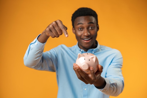 Young african amercian man holding piggy bank over yellow background