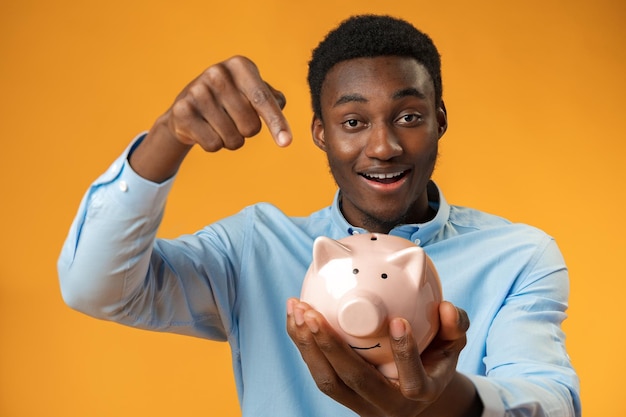 Young african amercian man holding piggy bank over yellow background