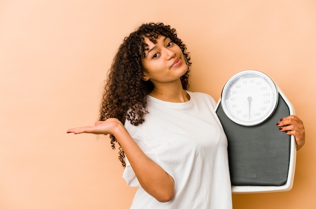 Young african american afro woman holding scales showing a copy space on a palm and holding another hand on waist.