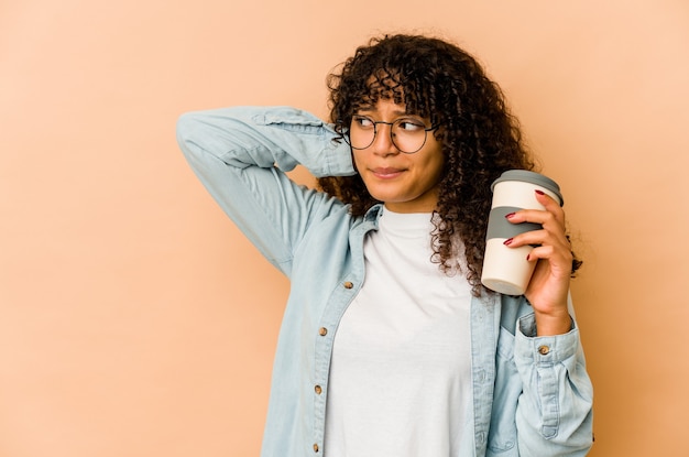 Giovane donna afroamericana afro che tiene un caffè da asporto che tocca la parte posteriore della testa, pensando e facendo una scelta.