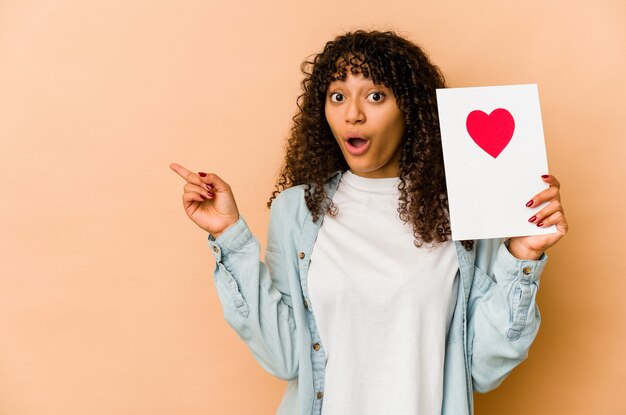 Young african american afro woman holding a valentines day card pointing to the side