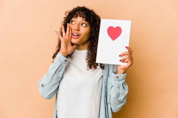 Young african american afro woman holding a valentines day card shouting and holding palm near opened mouth.