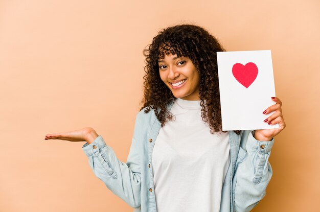 Young african american afro woman holding a valentines day card showing a copy space on a palm and holding another hand on waist.