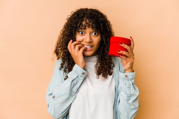 Young african american afro woman holding a valentines day gift biting fingernails, nervous and very anxious.