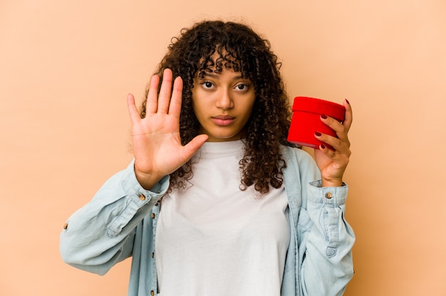 Young african american afro woman holding a valentines day gift standing with outstretched hand showing stop sign, preventing you.