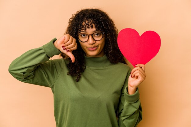 Giovane donna afroamericana afro che tiene un cuore di san valentino che mostra un gesto di avversione, pollice in giù. concetto di disaccordo.