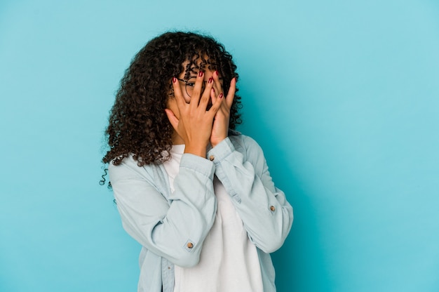 Young african american afro woman isolated blink at the camera through fingers, embarrassed covering face.