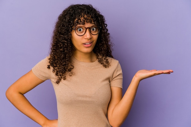 Young african american afro woman isolated showing a copy space on a palm and holding another hand on waist.