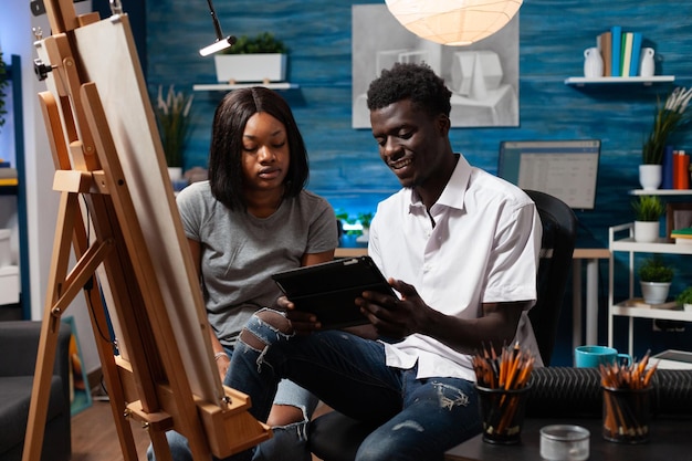 Young african american artistic partners watching video\
tutorials of drawing techniques on electronic tablet sitting in\
home art workshop. friends having good time learning new sketching\
abilities.