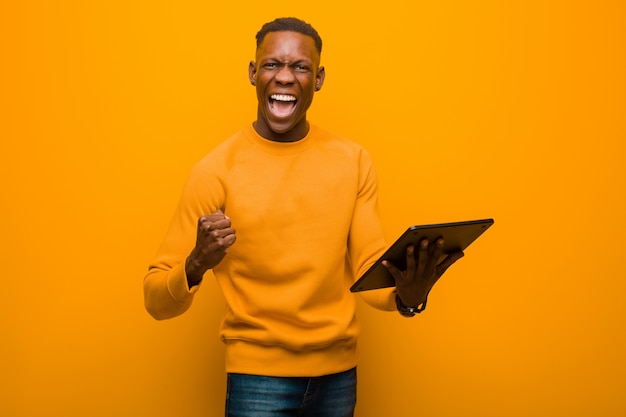 Young african american black man against orange wall with a smart tablet