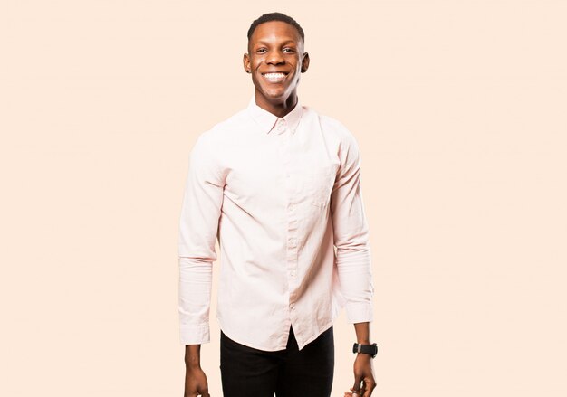 Young african american black man looking happy and goofy with a broad, fun, loony smile and eyes wide open against beige wall