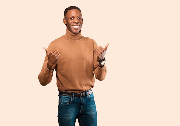 Young african american black man smiling joyfully and looking happy, feeling carefree and positive with both thumbs up against beige wall