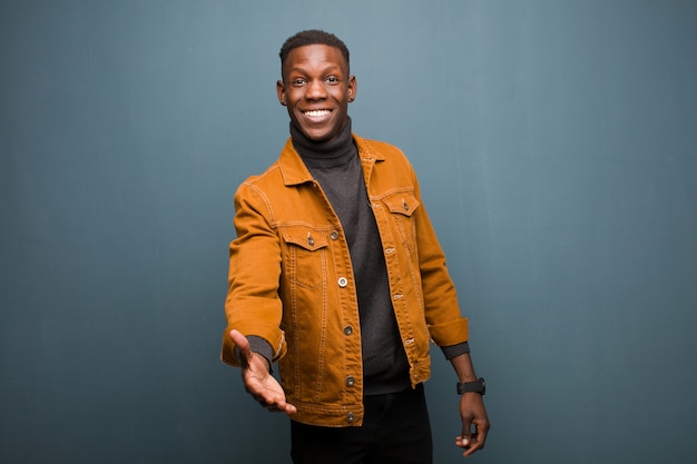 Young african american black man smiling, looking happy, confident and friendly, offering a handshake to close a deal, cooperating against grunge wall