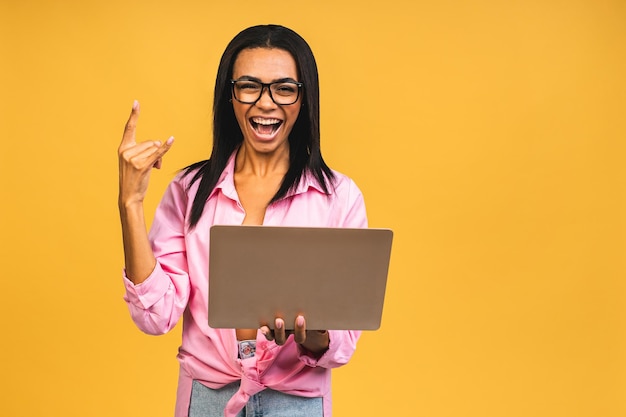 Young african american black positive cool lady with curly hair using laptop and smiling isolated over yellow background