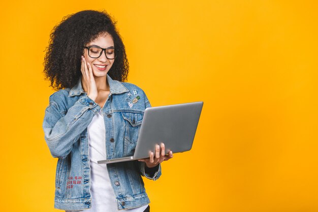 Young african american black positive cool lady with curly hair using laptop and smiling isolated over yellow background.