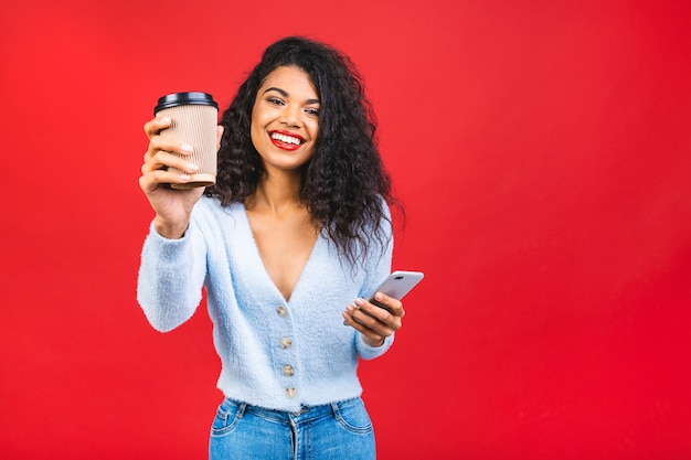 Giovane donna nera afroamericana che tiene una tazza di caffè e un telefono cellulare isolato su sfondo rosso