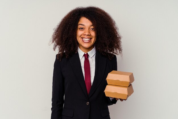 Молодая афро-американская бизнес-леди, держащая гамбургер на белом фоне, счастливая, улыбающаяся и веселая.