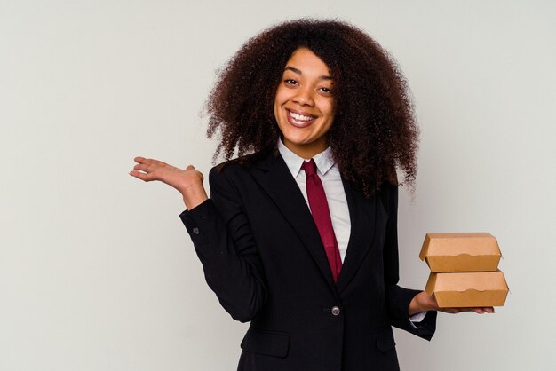 Young african american business woman holding a hamburger isolated on white wall showing a copy space on a palm and holding another hand on waist.