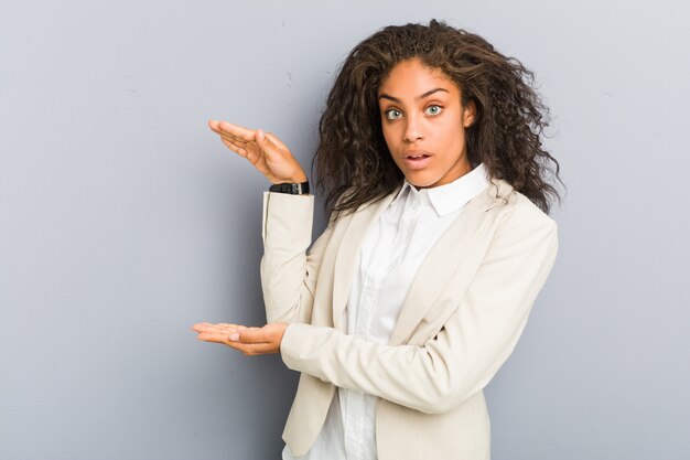 Young african american business woman shocked and amazed holding a copy space between hands.