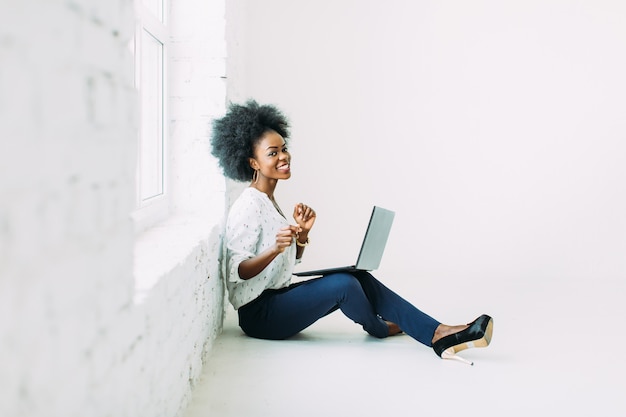 Молодой афроамериканец бизнес женщина, используя ноутбук, сидя на полу возле большого окна