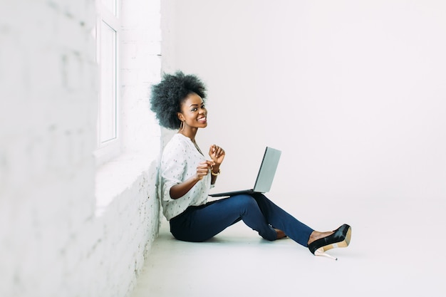 Young african american business woman using the laptop, while sitting on the floor near a big window